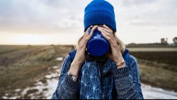 An older woman wearing a blue knit cap drinks coffee on a beach