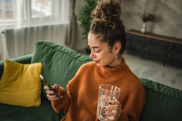 woman sitting on sofa drinking water looking at phone